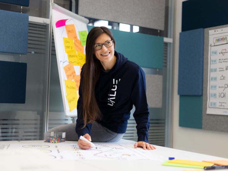 Ina Krasowski bei der Arbeit, sitzt lässig halb auf einem Tisch mit einem Edding in der Hand, vor sich ein großes Plakat, lächelt und schaut interessiert, im Hintergrund ein Flipchart mit Post Its